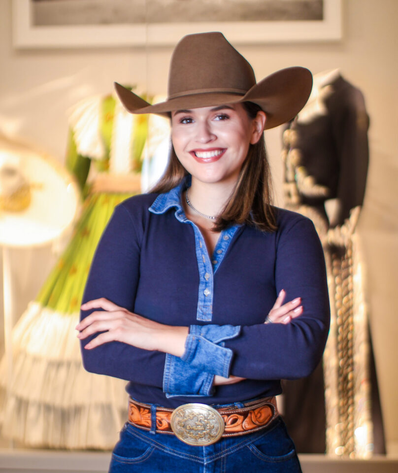 Woman smiling, wearing a brown cowboy hat, navy shirt with denim collar, and a large belt buckle, arms crossed.