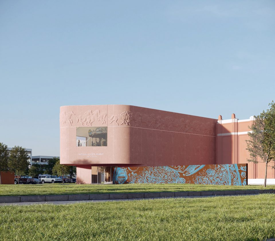 A modern pink building with a curved corner features sculpted reliefs and a large window. Colorful blue and orange patterns decorate the lower wall. Trees and grass surround the structure under a clear sky.
