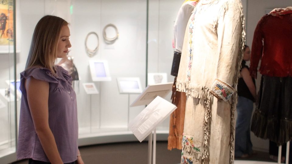 Woman in a purple blouse stands to the left, viewing a display of fringed dresses in a museum exhibit.
