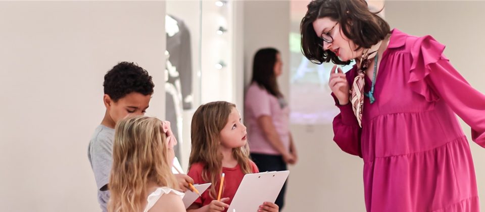 A woman in a magenta dress speaks to three children holding clipboards and pencils inside a brightly lit gallery; another adult stands in the background.