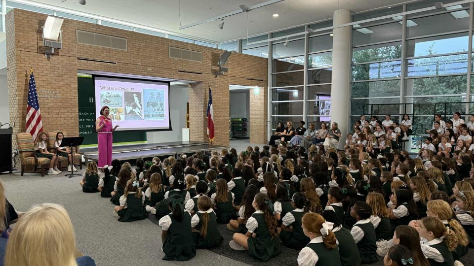 Teacher in pink dress speaks to a large group of students seated on the floor in a school auditorium; flags and a projection screen visible on stage.
