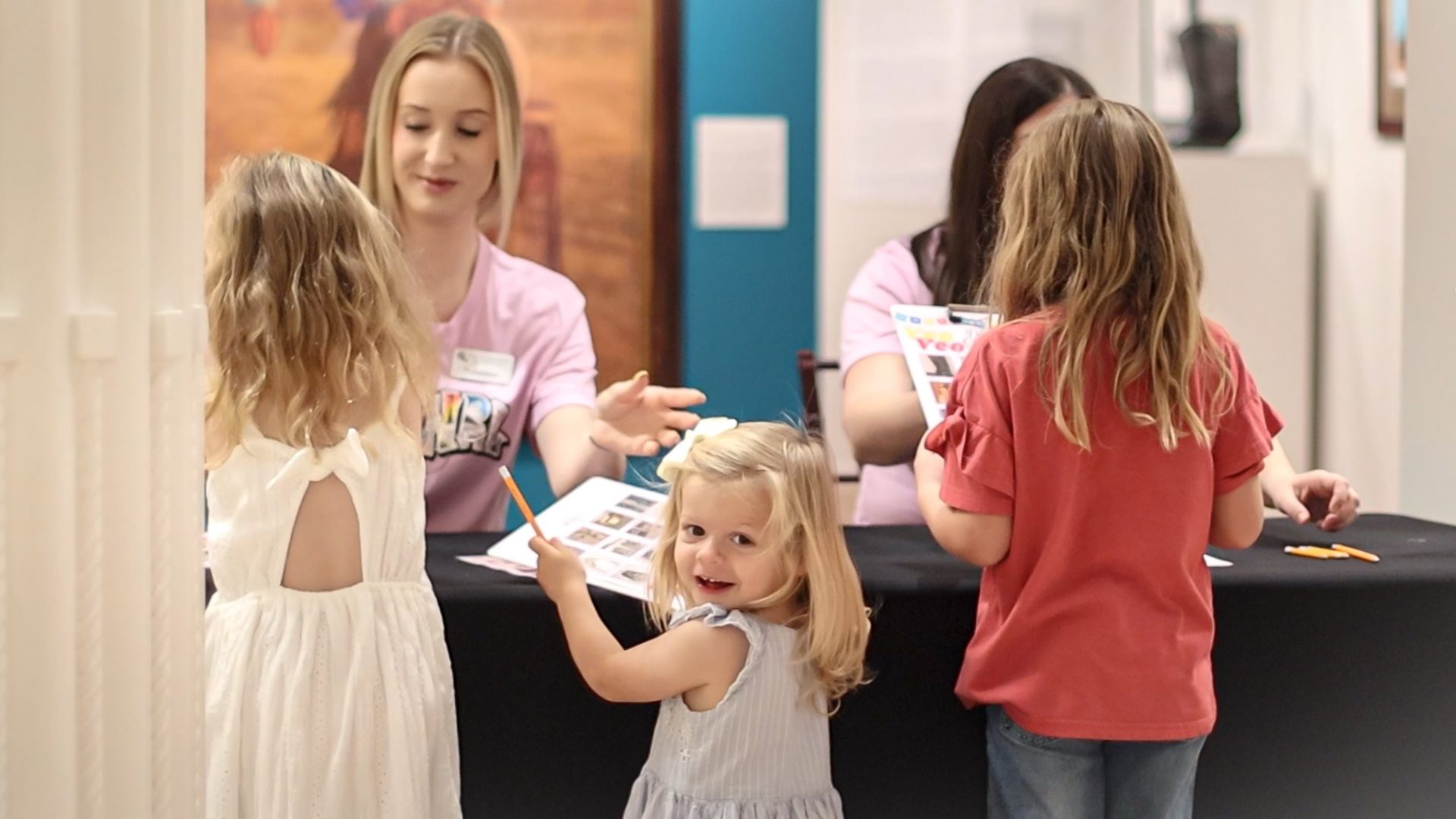 Young girl in a white dress smiles at the camera while staff assist children at a busy desk in a hallway.