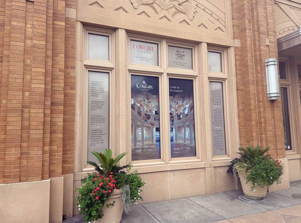 Brick facade with tall window displays showing Cowgirl Magazine posters and quotes, flanked by potted plants in the foreground.