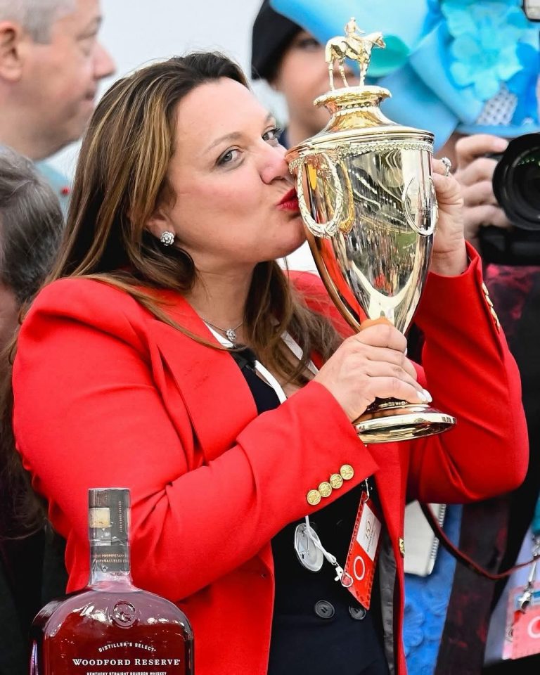 Woman in a red blazer kisses a large gold trophy at a celebration, with a bottle of Woodford Reserve visible in the foreground.