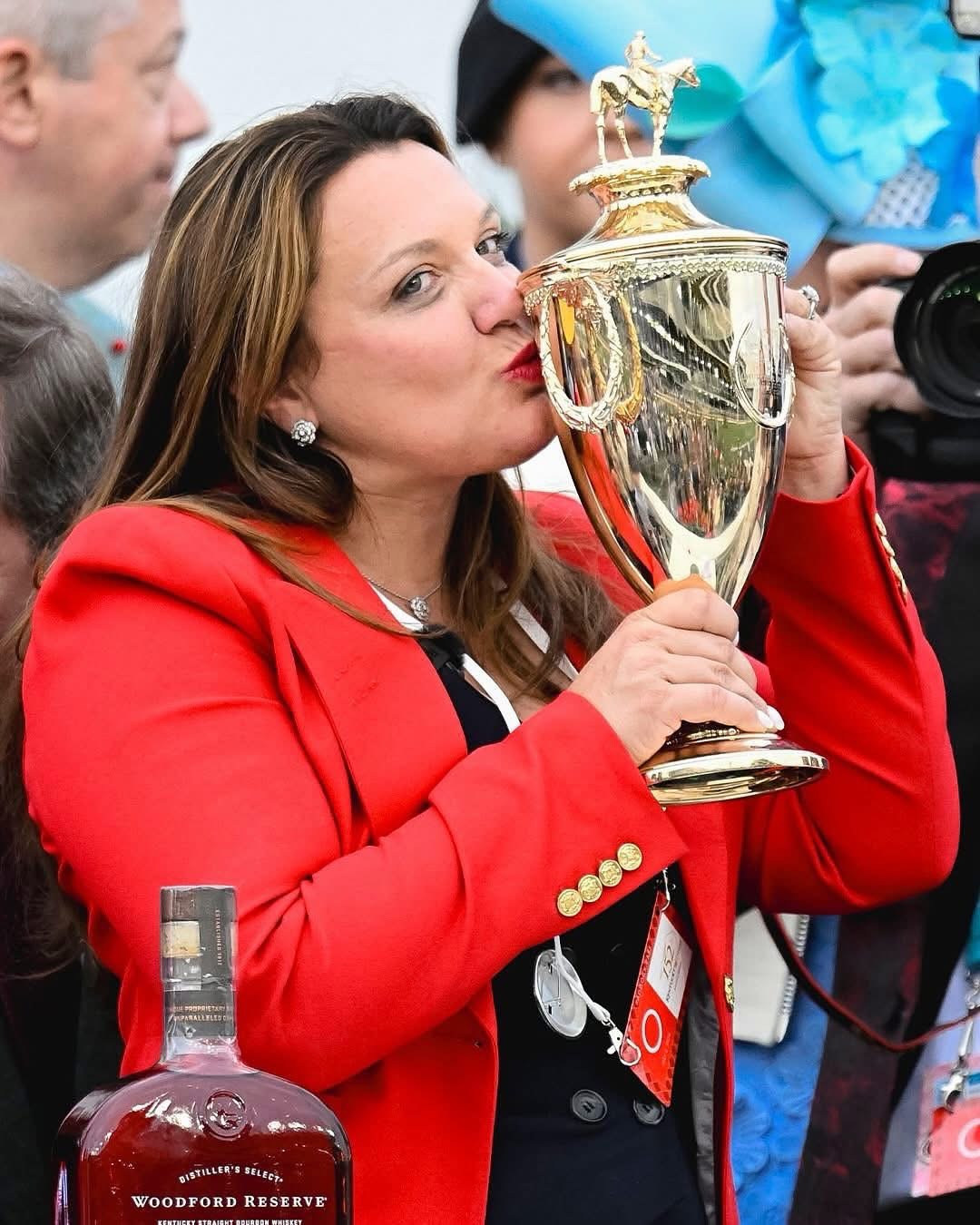 Woman in a red blazer kisses a large gold trophy at a celebration, with a bottle of Woodford Reserve visible in the foreground.