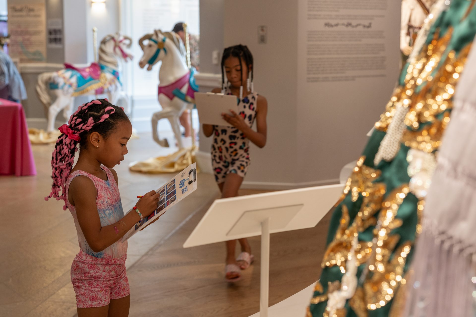 Young girl with pink braids reads a booklet in a museum exhibit with carousel horses in the background, suggesting a gallery or display area.