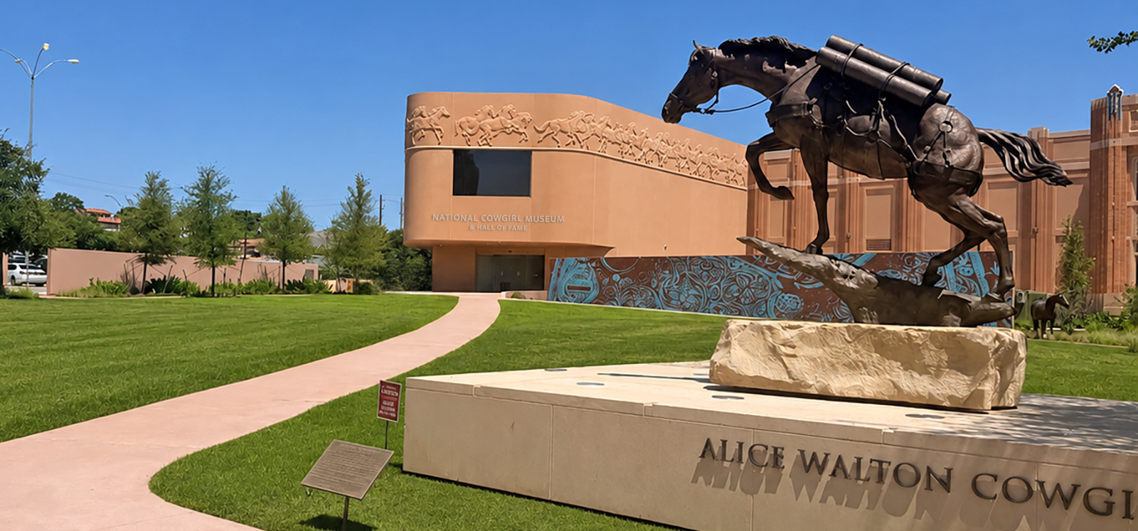 Bronze statue of a galloping horse on a stone pedestal in front of the National Cowgirl Museum and Hall of Fame building with a curved beige facade and horse-relief artwork, on a sunny day.