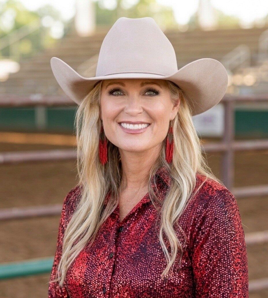 Smiling woman in a beige cowboy hat and red patterned shirt outdoors at a ranch, with long blonde hair and red earrings.