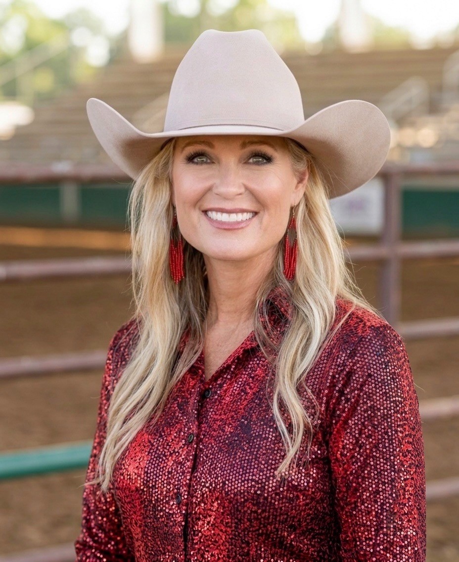 Smiling woman in a beige cowboy hat and red patterned shirt outdoors at a ranch, with long blonde hair and red earrings.