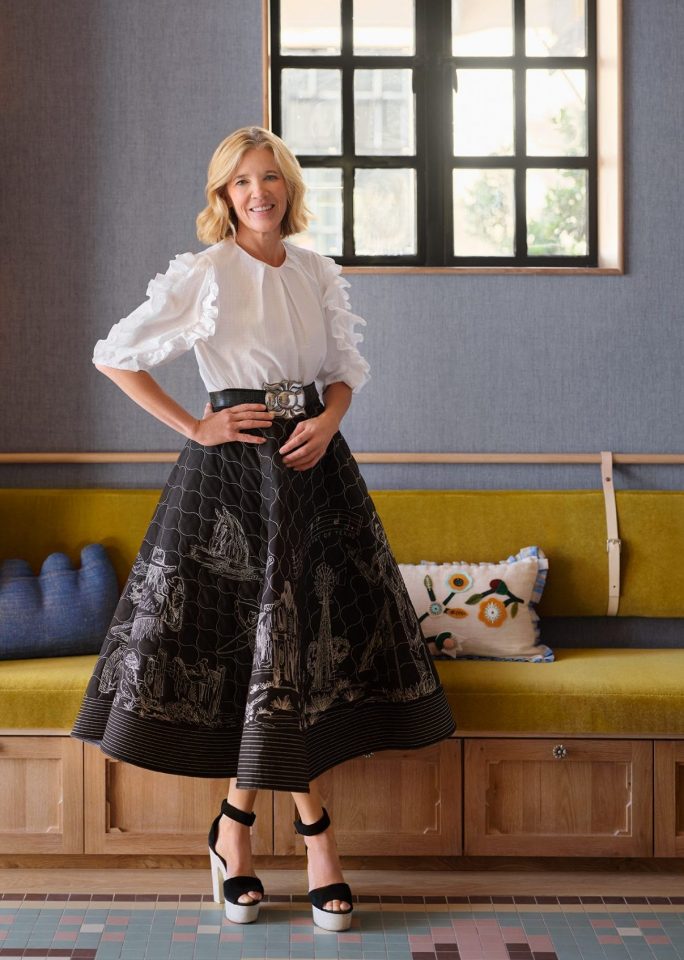 Smiling woman in a white blouse and black patterned skirt stands in a wood-framed window alcove with a yellow bench beneath.