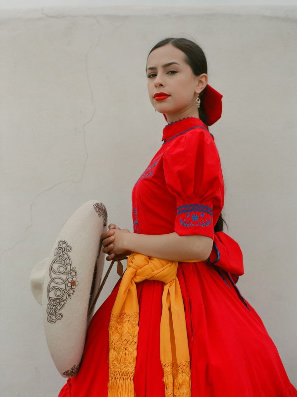 Woman in a bright red traditional dress with blue embroidery, standing against a pale wall, holding a decorative hat with a yellow sash.