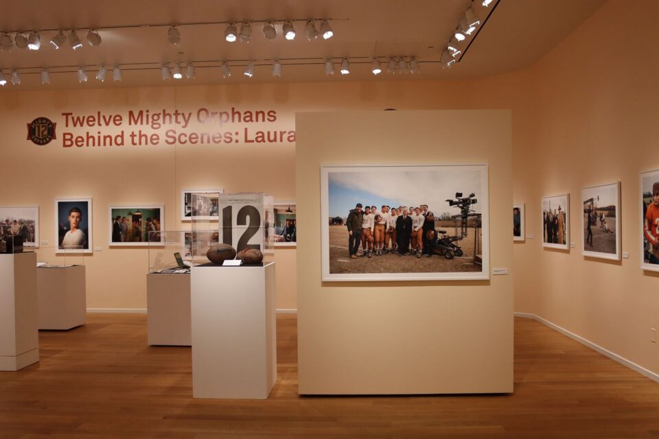 Museum gallery with wall text reading 'Twelve Mighty Orphans Behind the Scenes: Laura' and framed photos along beige walls