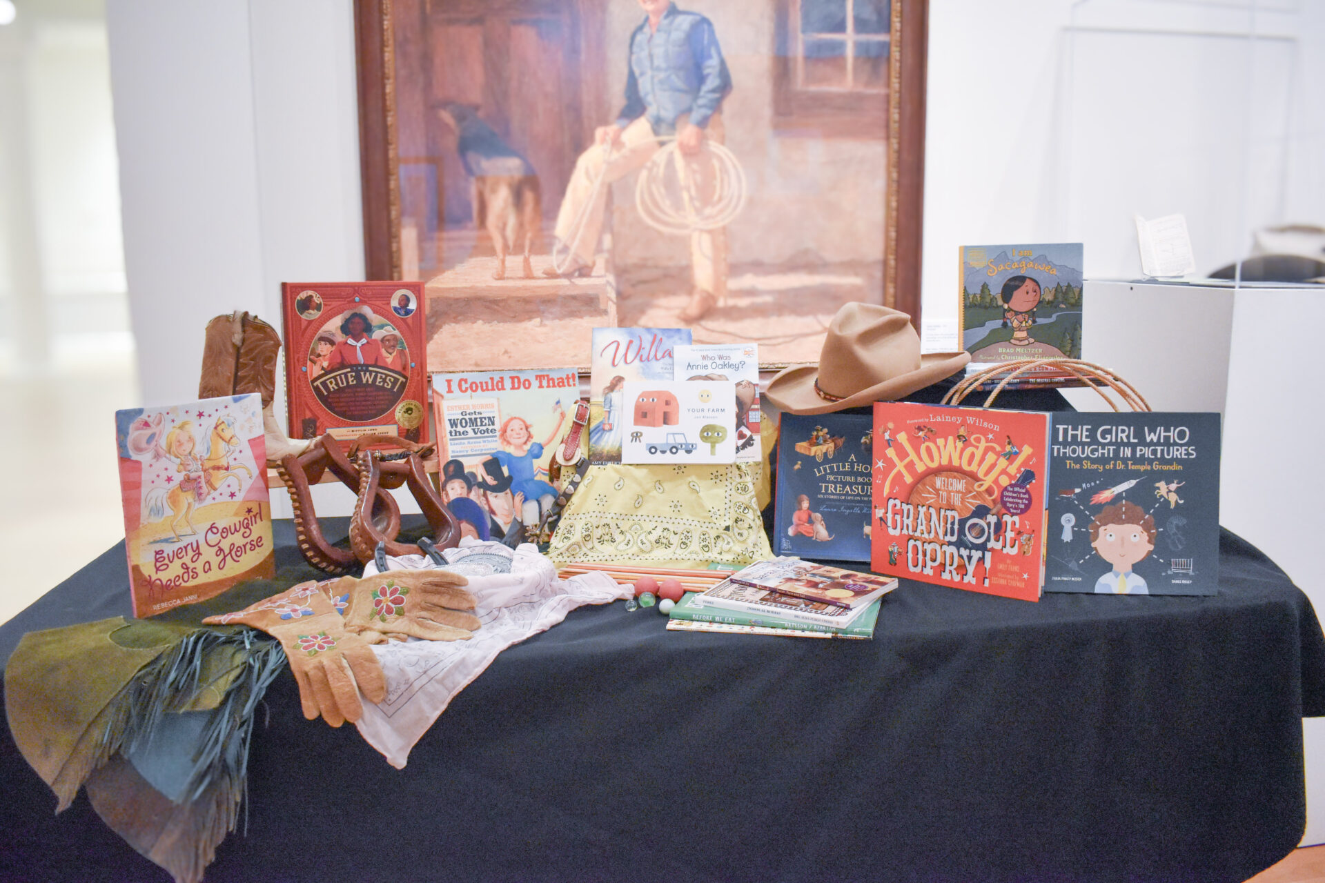 Display table of vintage children's books and western-era props arranged in front of a large painting in a museum exhibit.