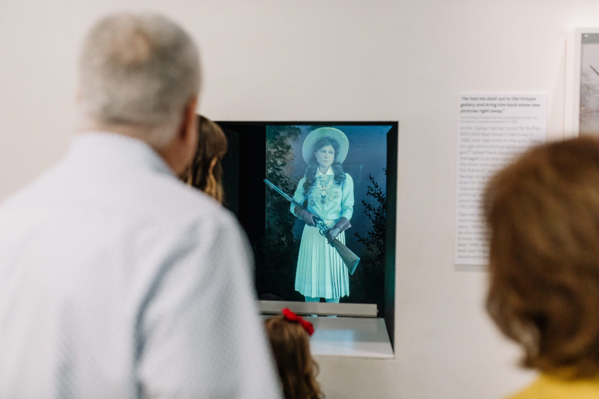 Two adults and a child observe a museum display showing a hologram of a young woman in old-fashioned attire holding a rifle, with informative text visible on the wall nearby.