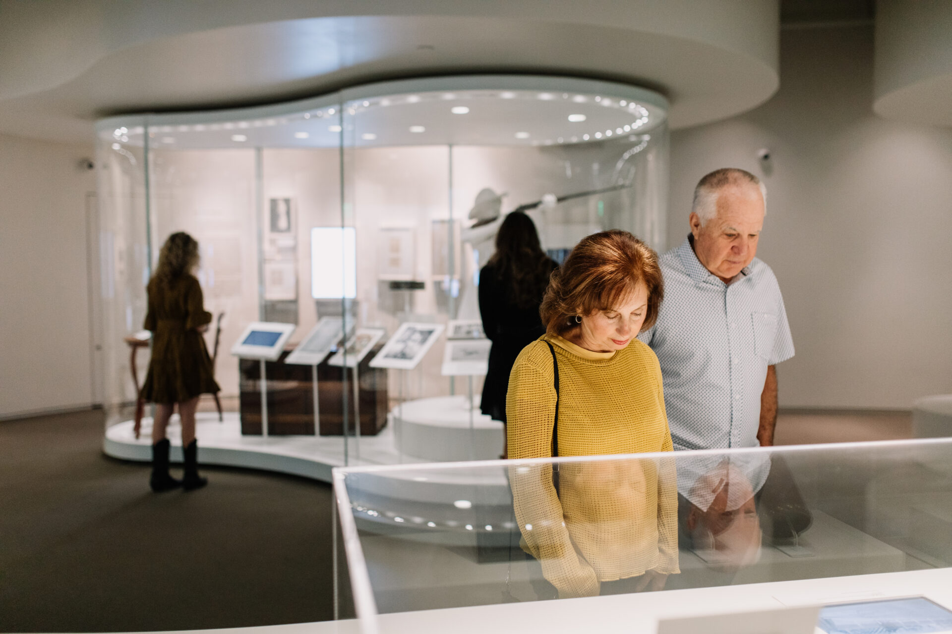 Two people view a display case in a modern, well-lit museum exhibit. Other visitors are visible in the background, looking at various displays and framed items inside glass cases.