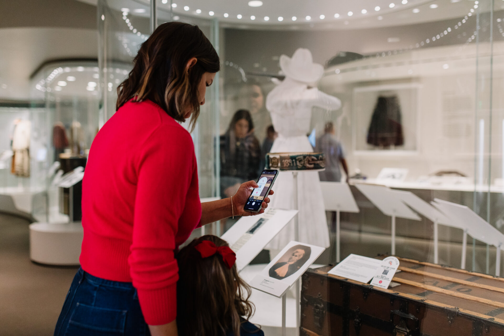 A woman in a red sweater uses her phone in a museum, standing beside a child. They view exhibits including a white dress and a vintage trunk displayed in glass cases.