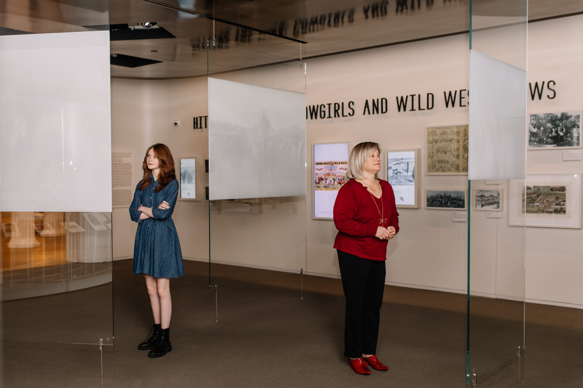 Two women stand apart in a museum exhibit with glass displays. One wears a blue dress with black boots and has arms crossed; the other wears a red top with black pants and red shoes. Historical photos are on the wall behind them.