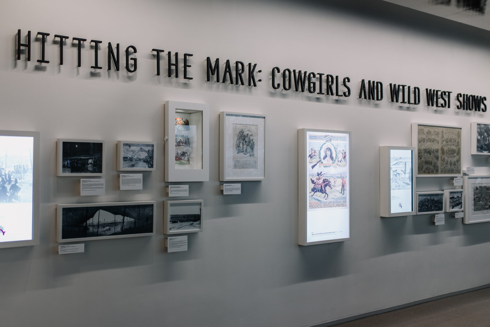 A museum wall display titled Hitting the Mark: Cowgirls and Wild West Shows features photos, documents, and posters, some in illuminated frames.