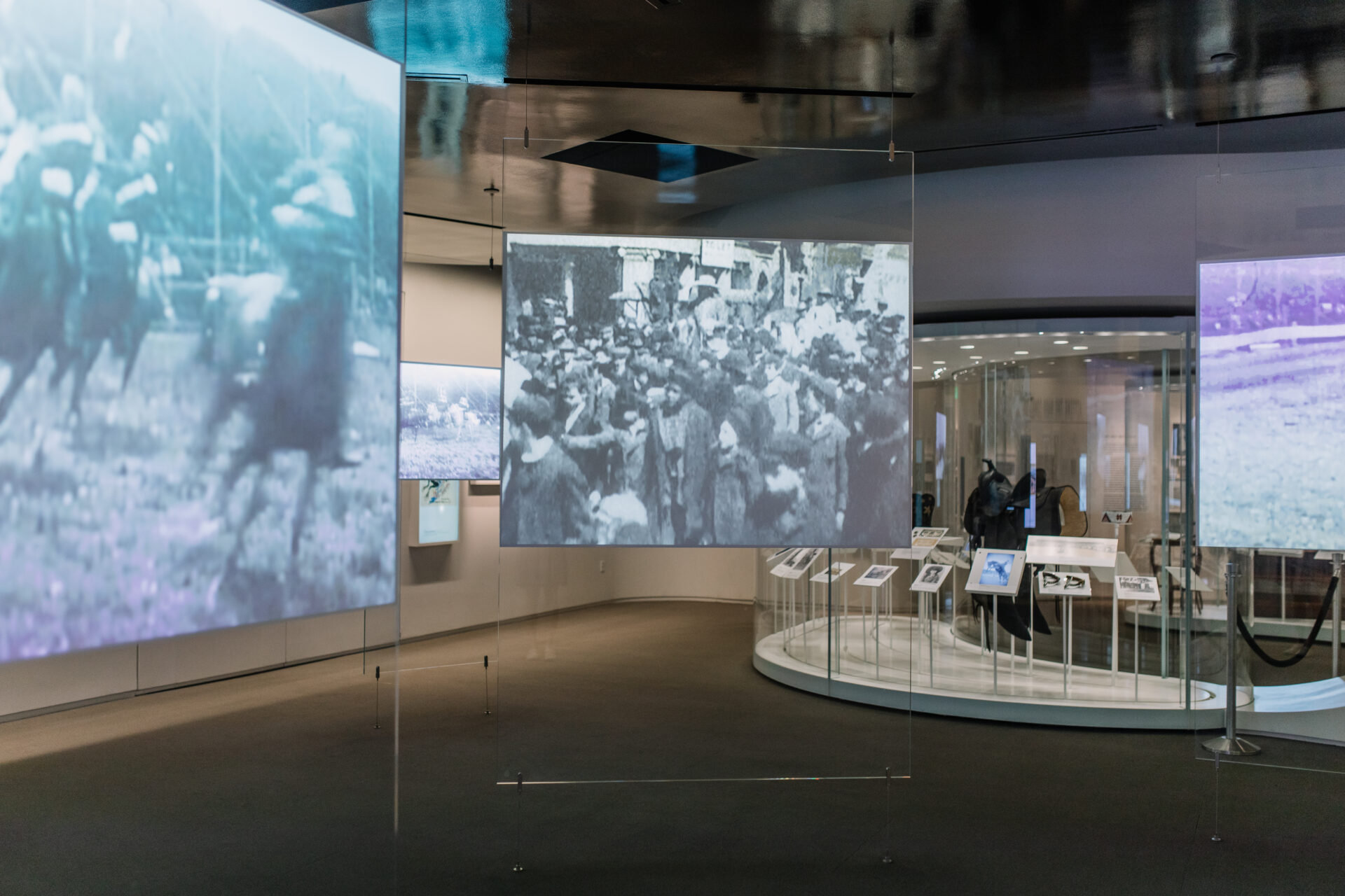 Museum exhibit showing suspended panels with historical black-and-white photographs and film projections. Display cases with artifacts and information screens are visible in the background under soft lighting.