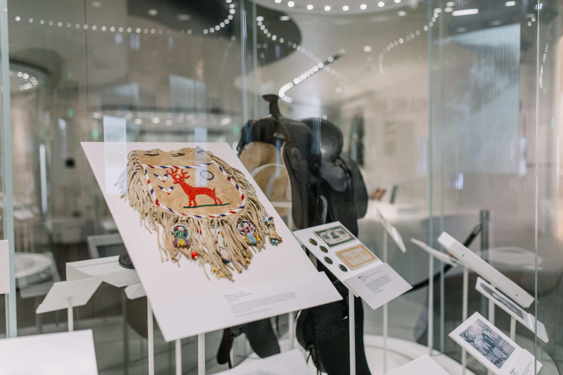 A glass display case features a fringed garment with a red deer design and colorful decorations, along with various artifacts and informational cards in a museum setting.