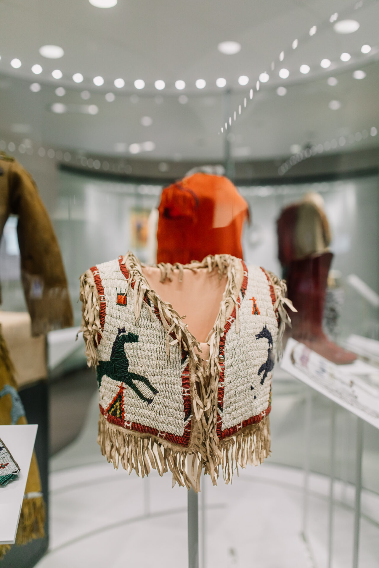 A beaded vest with fringes, featuring horse designs and colorful patterns, is displayed in a glass museum case with other artifacts visible in the background.
