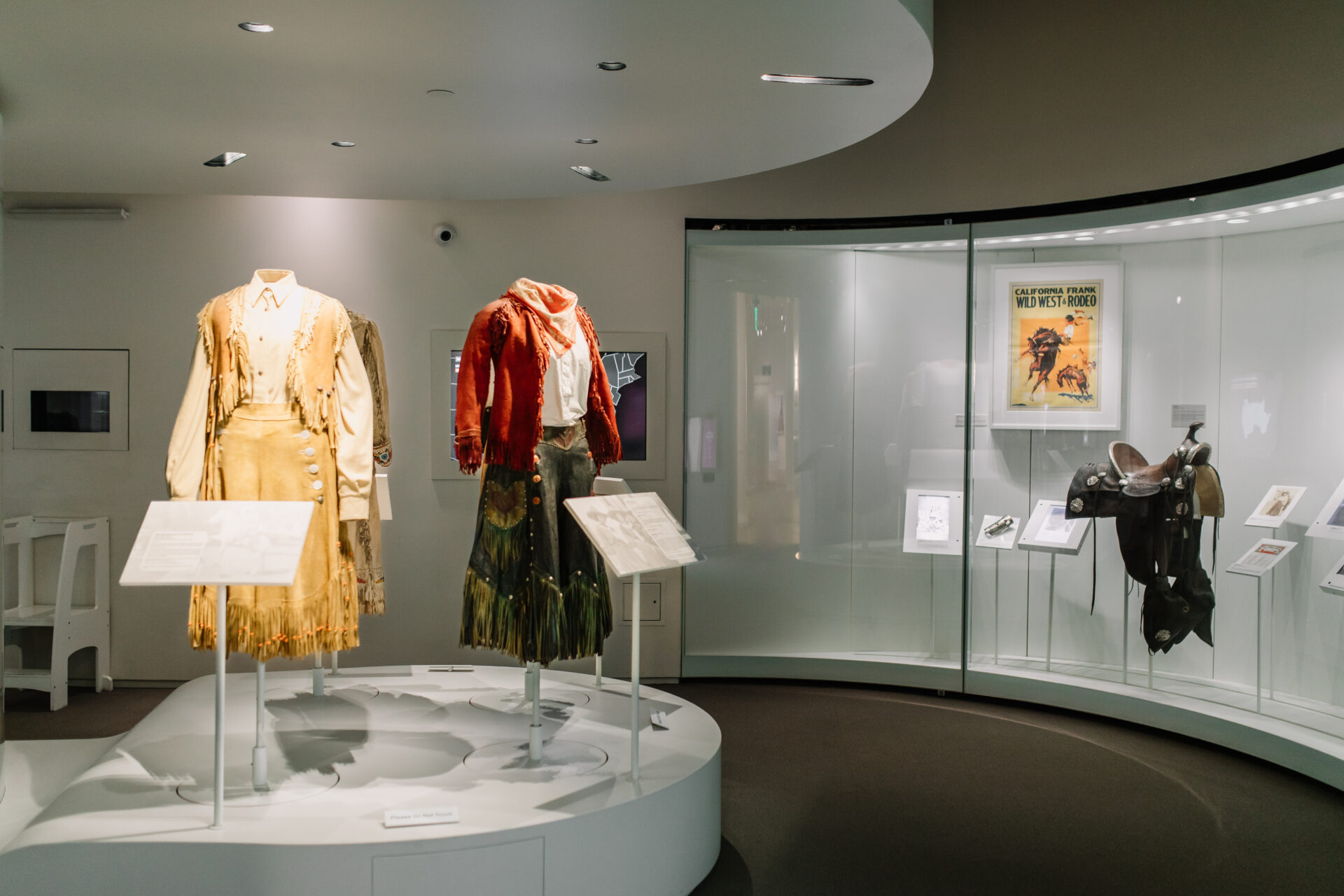 Museum display with two mannequins dressed in Western attire, a saddle, and various framed posters and documents in a glass case. The room is well-lit with modern white walls and display stands.
