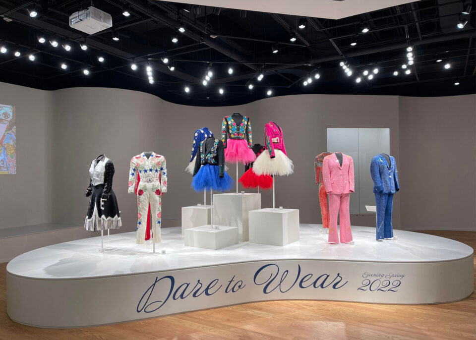 Colorful fashion costumes on mannequins displayed on a circular white platform in a gallery, with bright overhead lights and the words 'Dare to Wear' visible on the front edge.