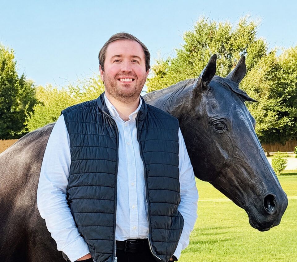 Man wearing a blue quilted vest and light shirt standing beside a black horse in a sunny grassy field.