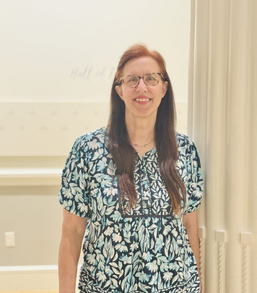 Smiling woman with brown hair and glasses in a blue floral blouse standing in a bright hallway.