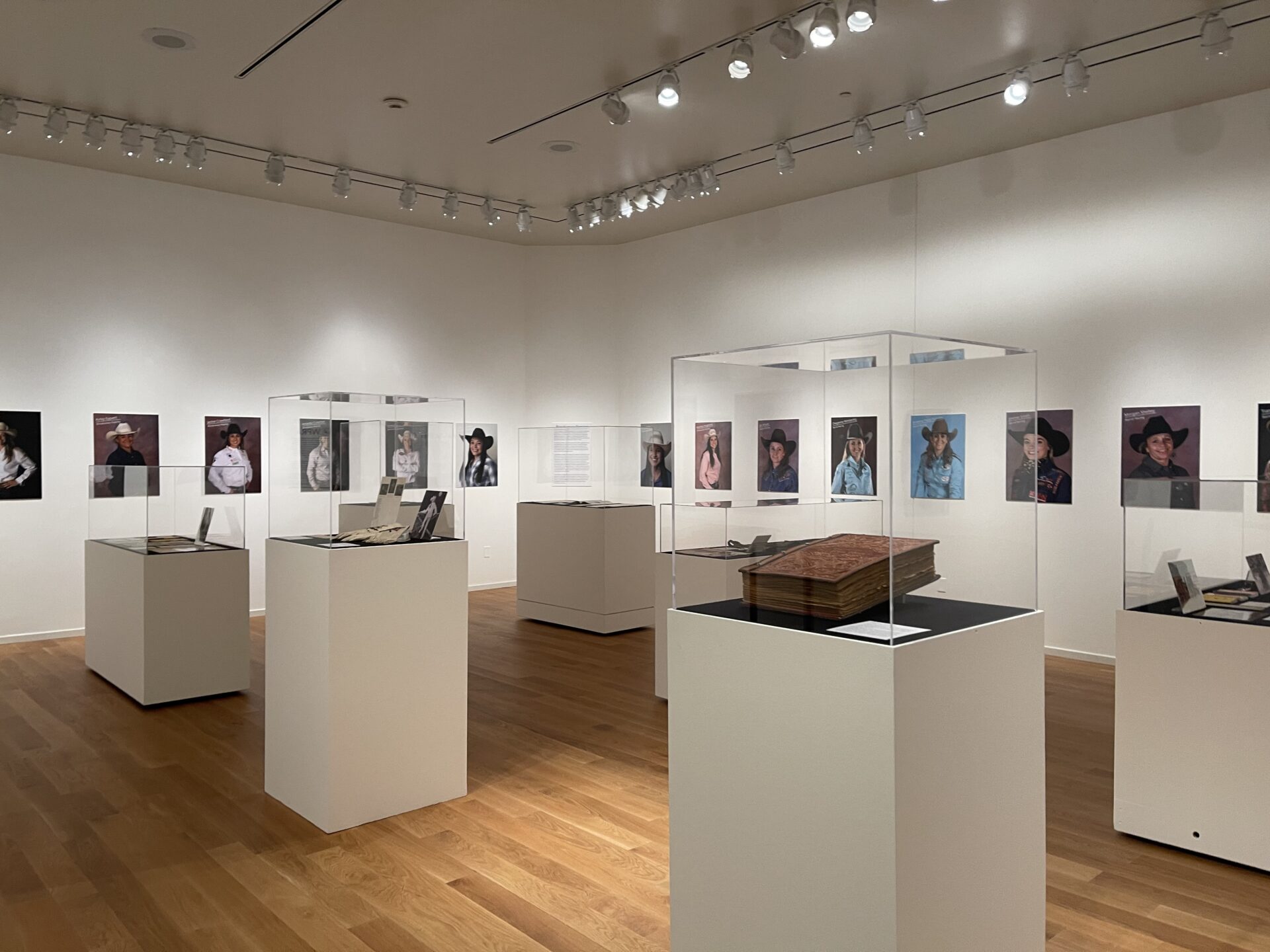 Museum gallery with framed portraits of people wearing hats along white walls, glass display cases, and a wooden floor. Lighting tracks illuminate the space.