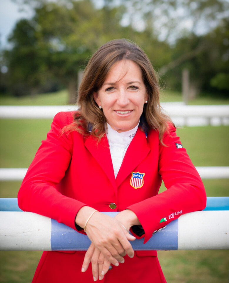 Woman in a red blazer leaning on a white-and-blue railing outdoors, smiling at the camera with a U.S.SA emblem on the blazer area (sports setting).