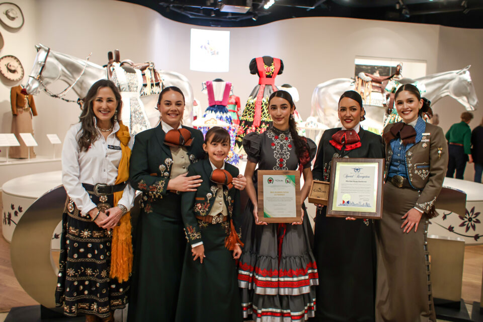 Group of women and a girl in ornate traditional dresses posing with certificates at an exhibition. A decorative horse sculpture is in the background.