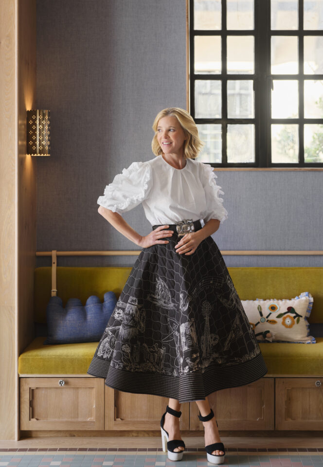 A woman in a white blouse and a black skirt with white designs stands smiling by a window, resting one hand on her hip in a stylish, modern room with yellow seating and decorative pillows.