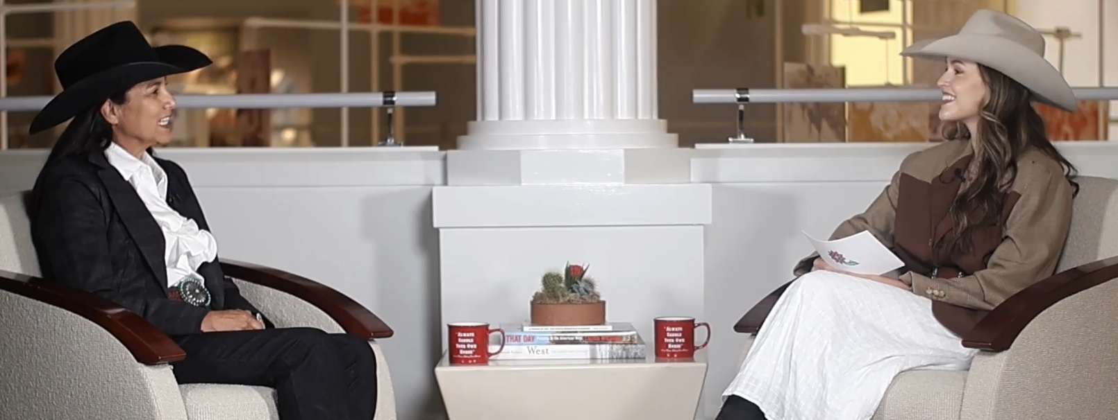 Two women in cowboy hats sit in beige armchairs across from each other in a modern lounge, with a small table between them holding a plant, magazines, and red mugs.
