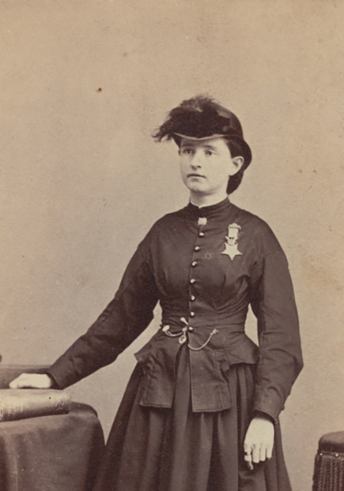 Victorian-era woman in a dark military-style dress with medal, standing beside a table.
