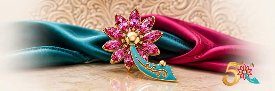 A gold and pink jeweled flower brooch with teal accents rests on marble, draped in teal and magenta silk. A gold 50th anniversary emblem with a similar flower appears in the corner.