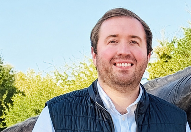 A man with short brown hair and a beard smiles outdoors, wearing a black vest over a white shirt, with green trees and a clear blue sky in the background.