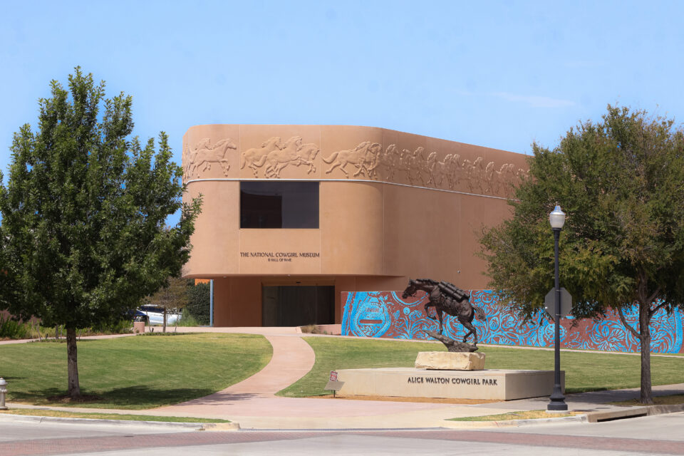 The National Cowboy Museum with tan curved walls, horse and rider reliefs, blue mural, green lawn, trees, and a bronze horse statue labeled Ailich Watson Cowgirl Park in front.