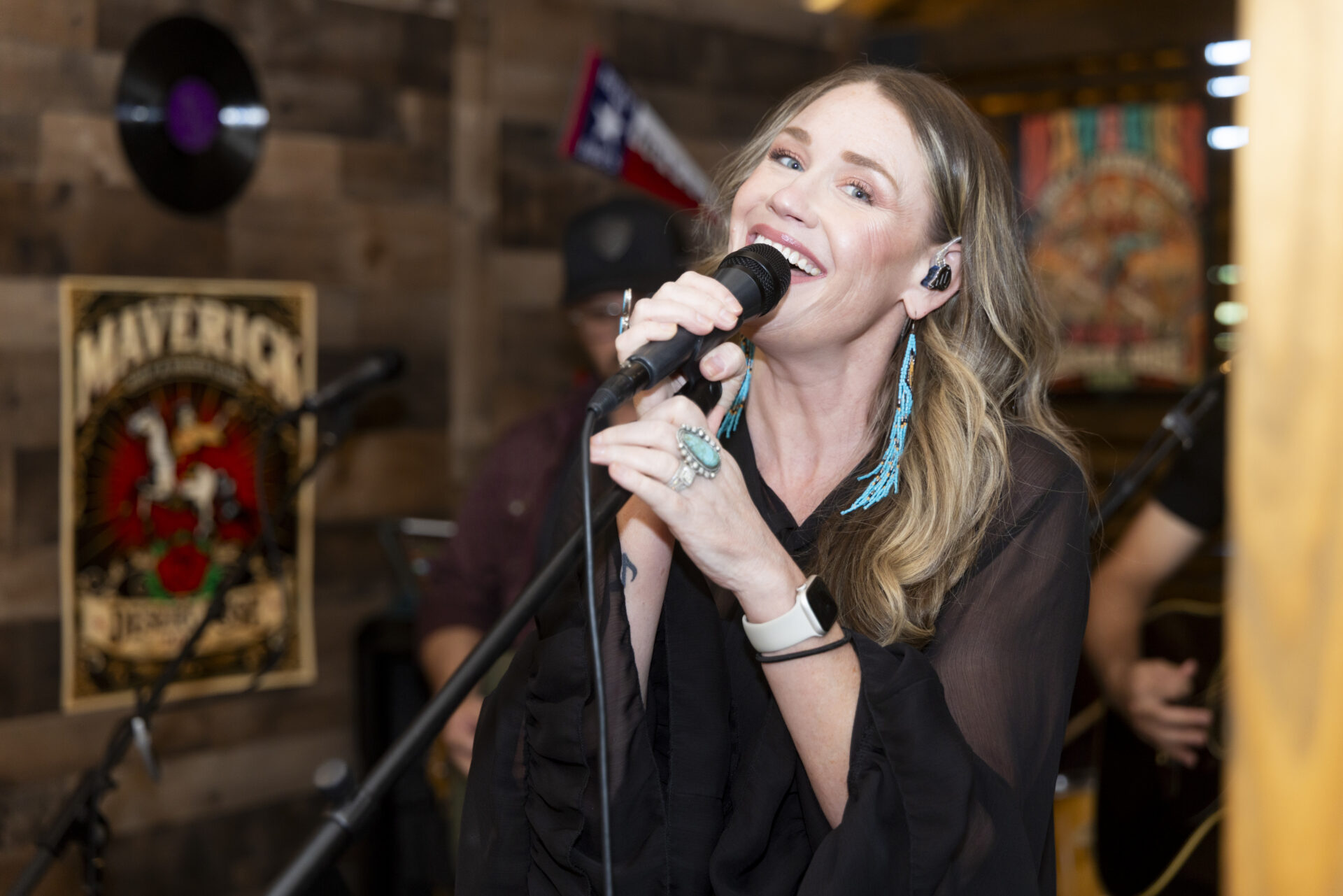 A female singer performing into a microphone on stage, with a band member visible in the background.