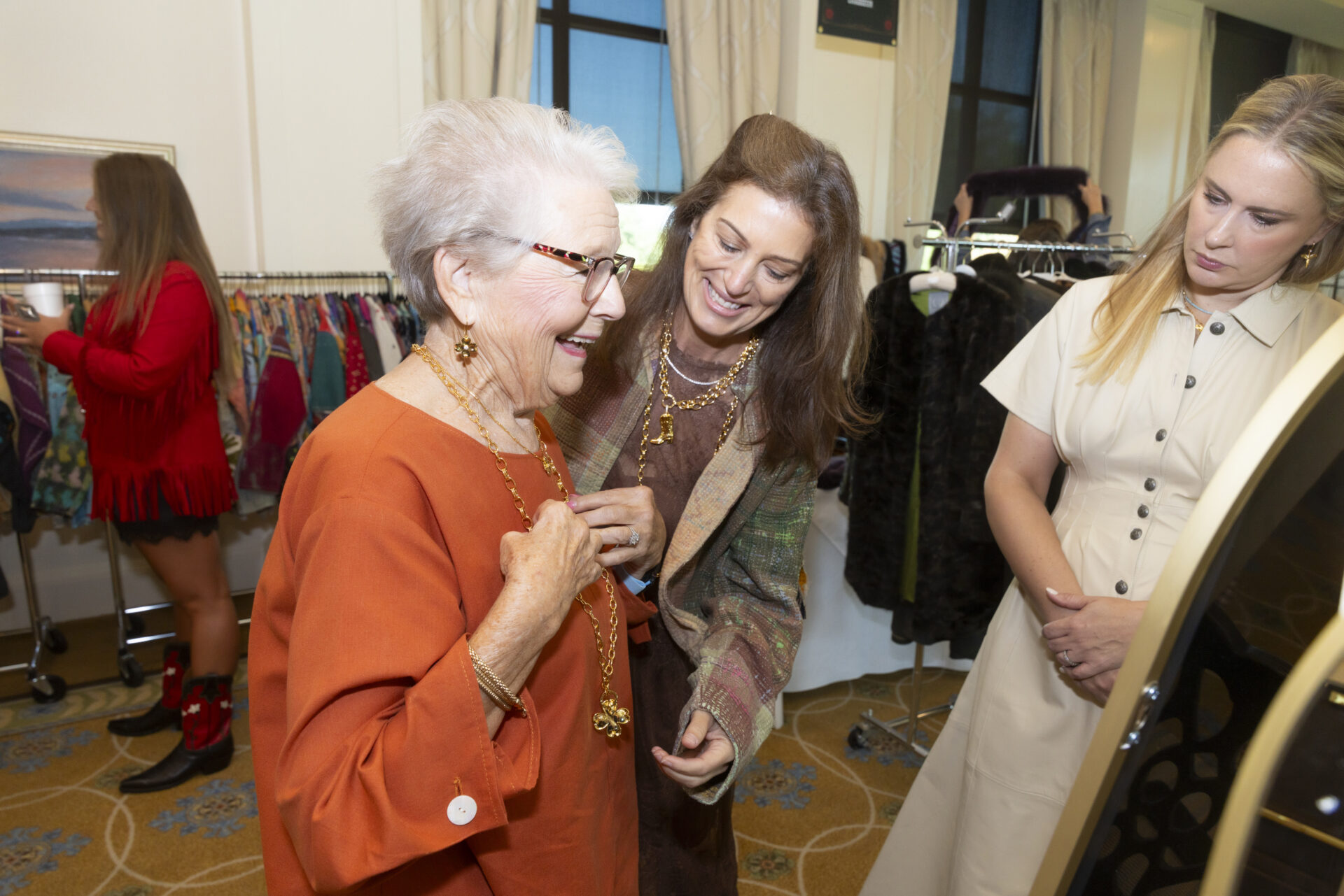 Three women interact at a clothing boutique event, with an older woman in an orange top smiling as she looks at a gold necklace.