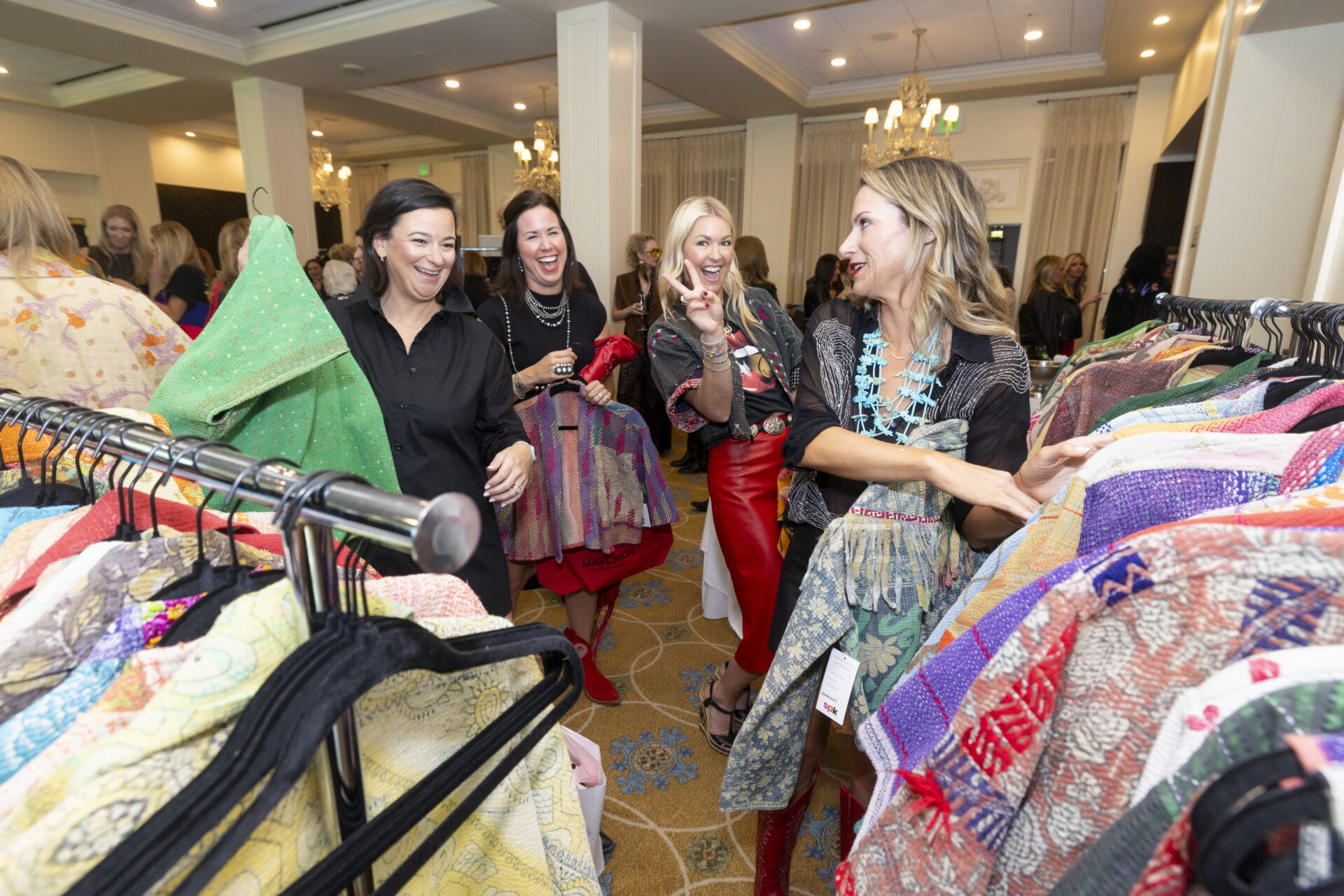 Four women smiling and laughing while browsing through colorful patterned jackets on clothing racks at an indoor event.