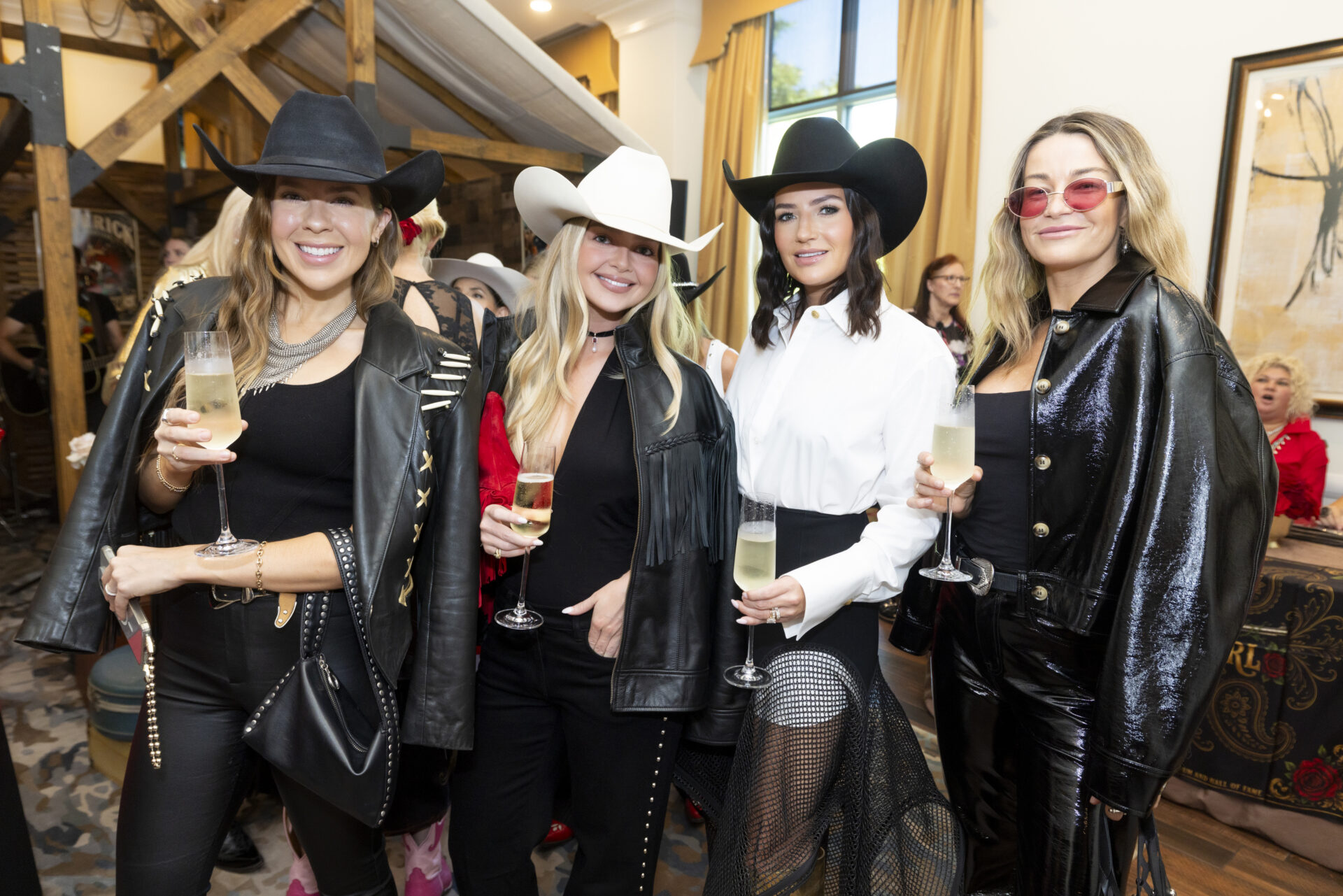 Four women wearing cowboy hats and stylish outfits pose together while holding glasses of champagne at an event.