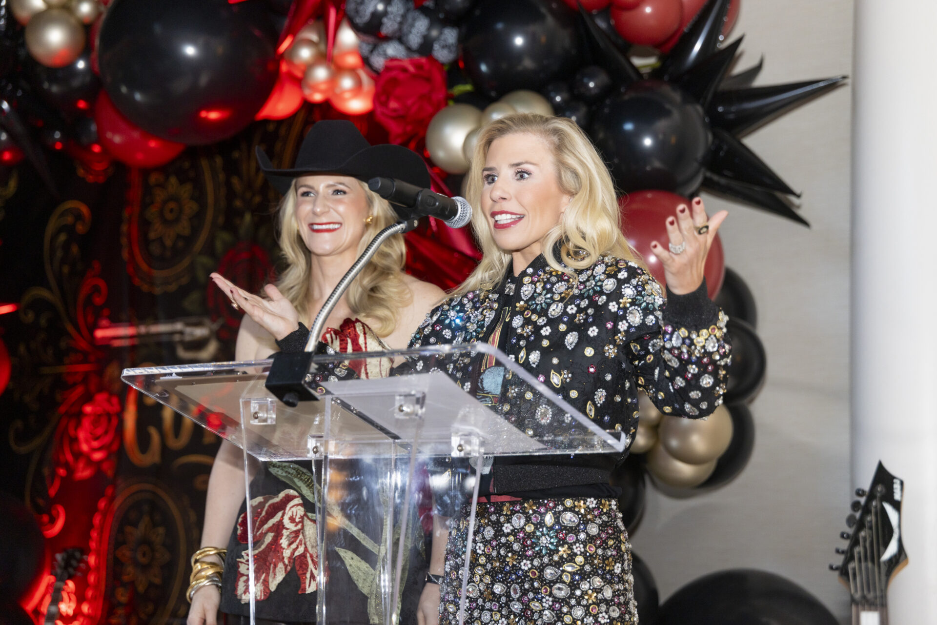 Two women stand behind a clear podium at an event, with one speaking into a microphone in front of a backdrop of black and red balloons.