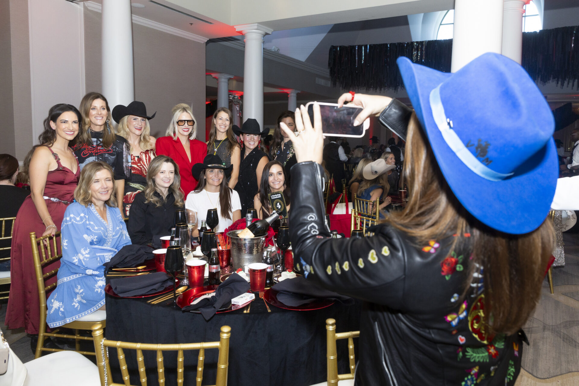 A person in a blue cowboy hat takes a photo of a group of women seated around a round dining table.