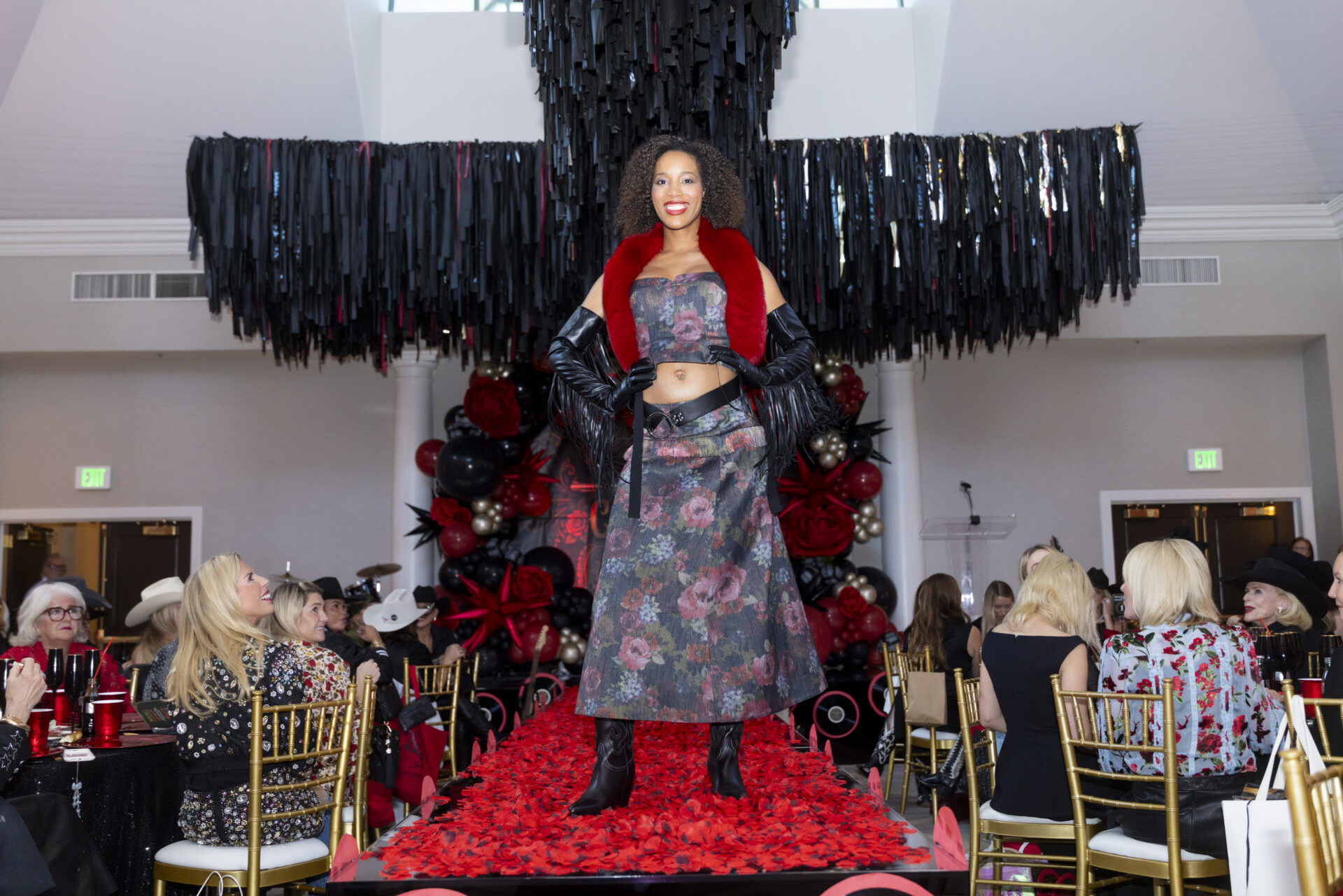 A model in a floral two-piece outfit with a red faux fur collar and black opera gloves walks a runway covered in red rose petals before a seated audience.