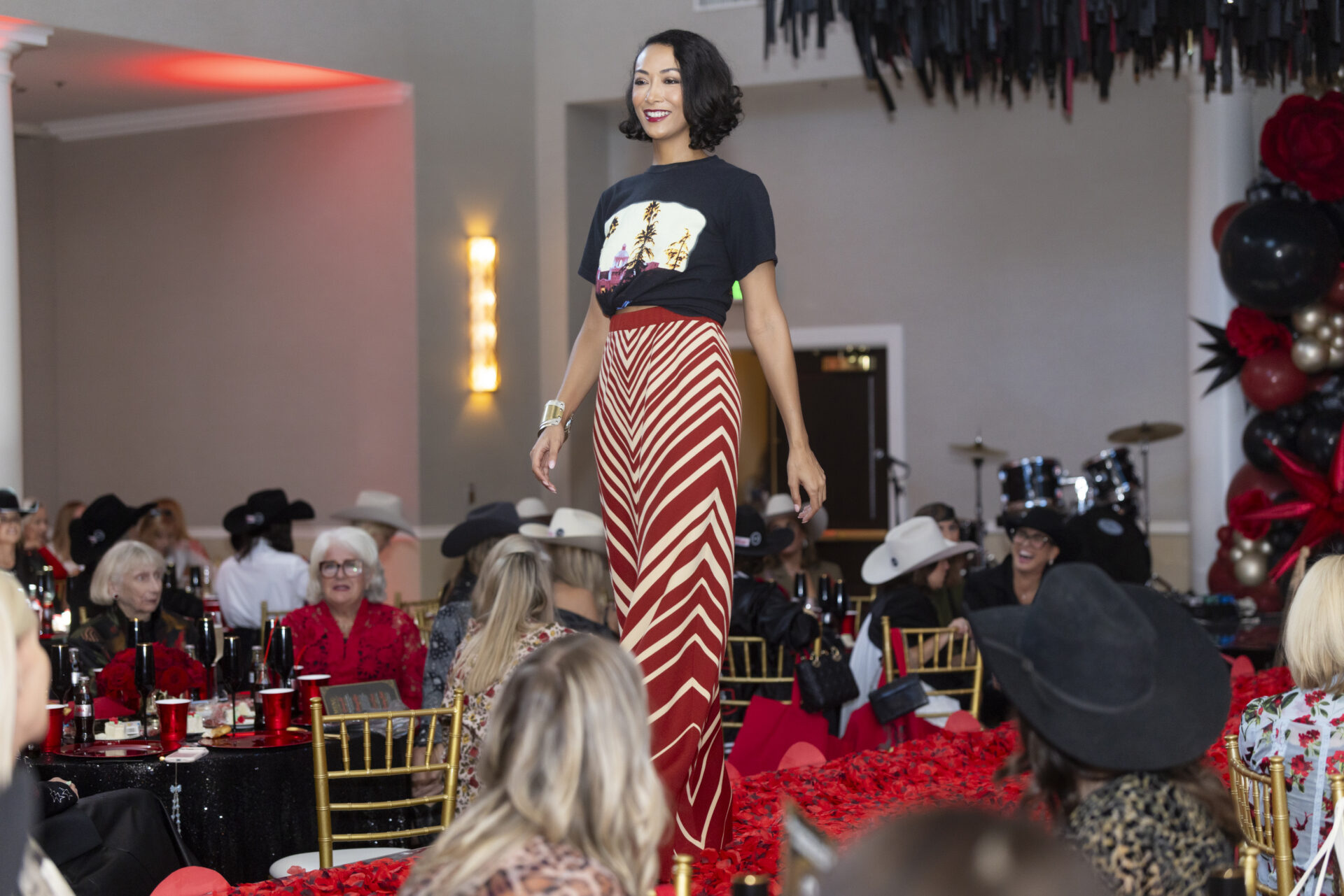 A woman with dark hair walks down a runway wearing a black t-shirt and a long, red and white chevron-patterned skirt, with audience members seated at tables nearby.