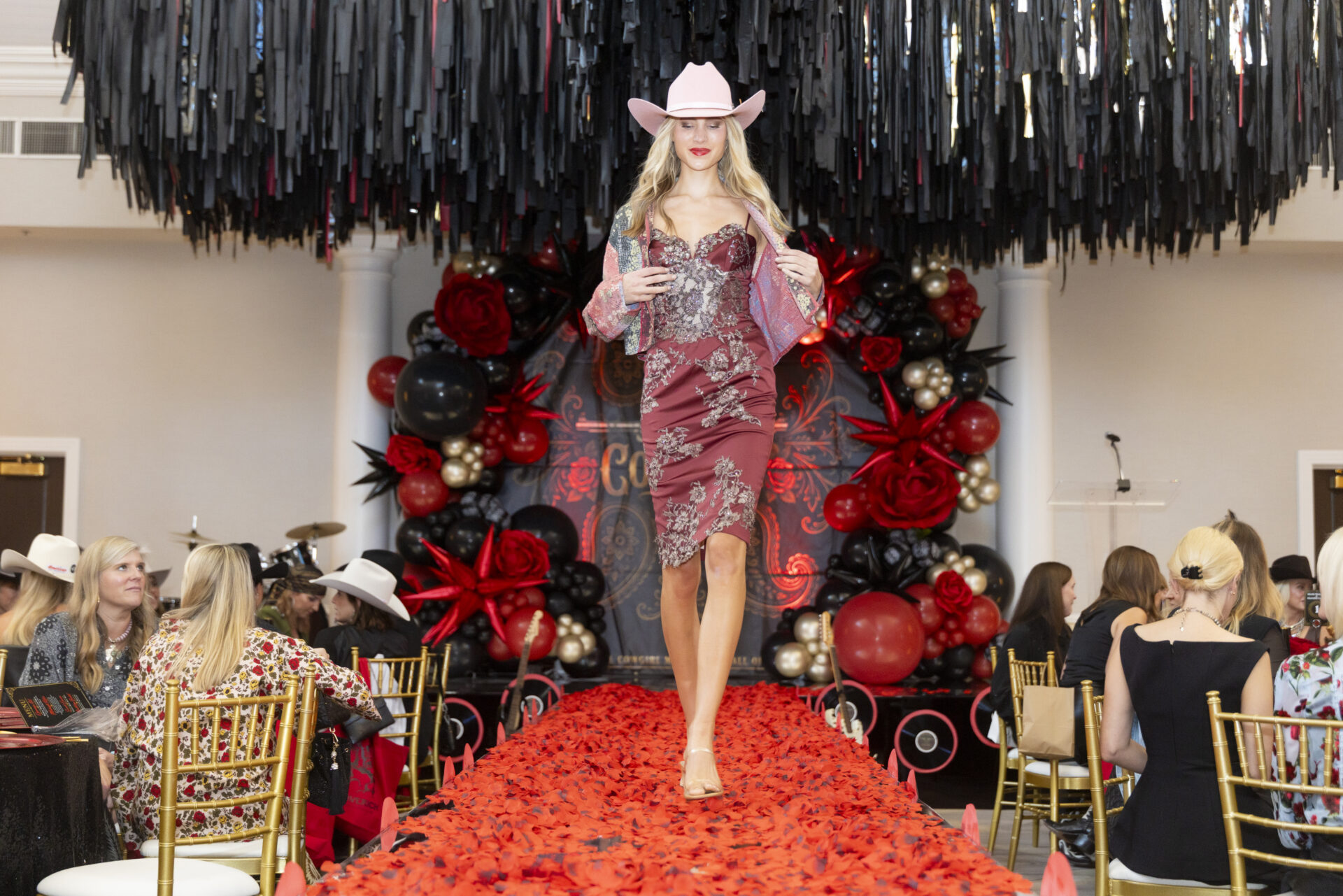 A model in a pink cowboy hat and embroidered maroon dress walks down a red petal-covered runway in front of a black and red balloon arch.