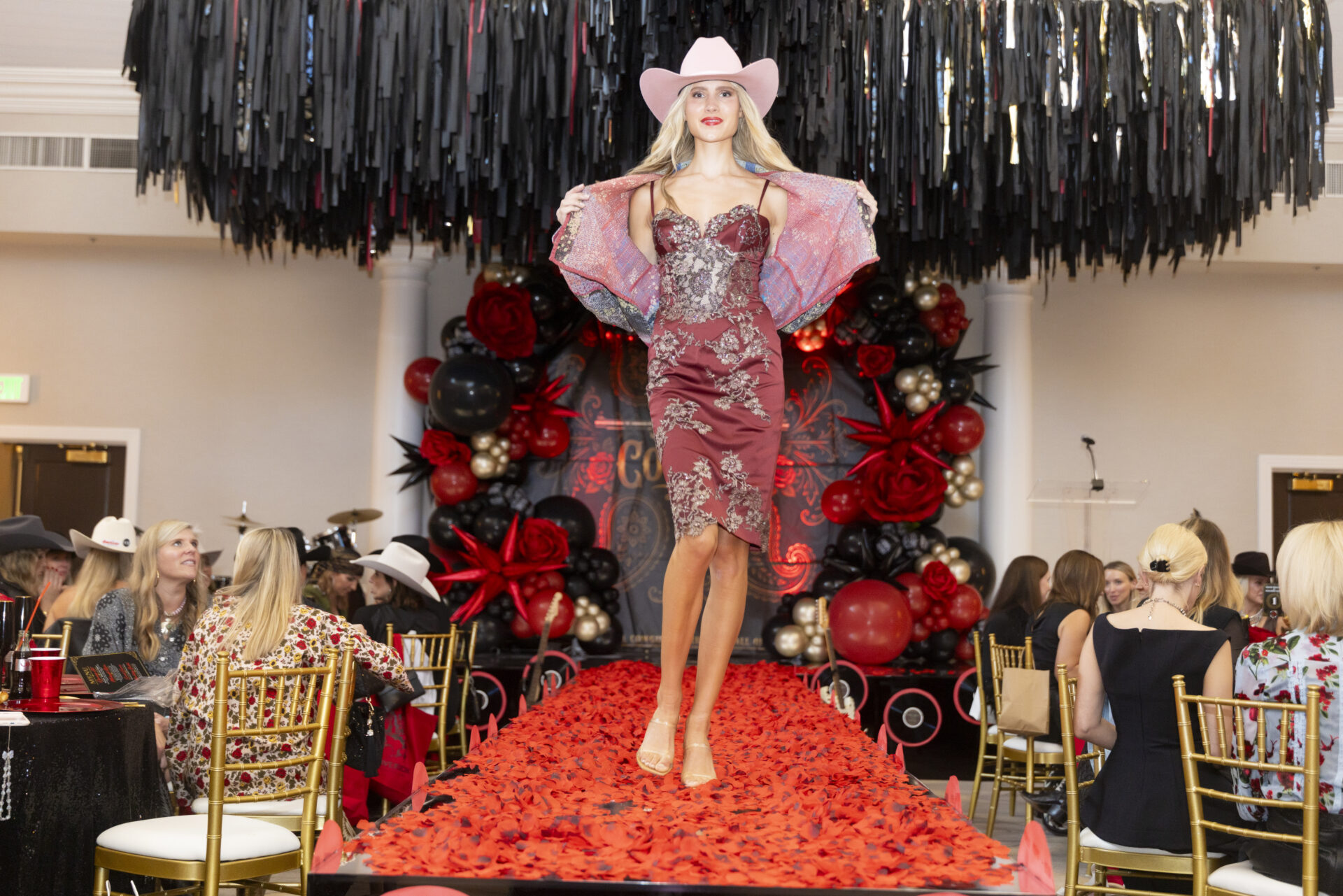 A model wearing a pink cowboy hat and a maroon, floral-embroidered dress walks down a runway covered in red rose petals, with an audience seated at tables on both sides.